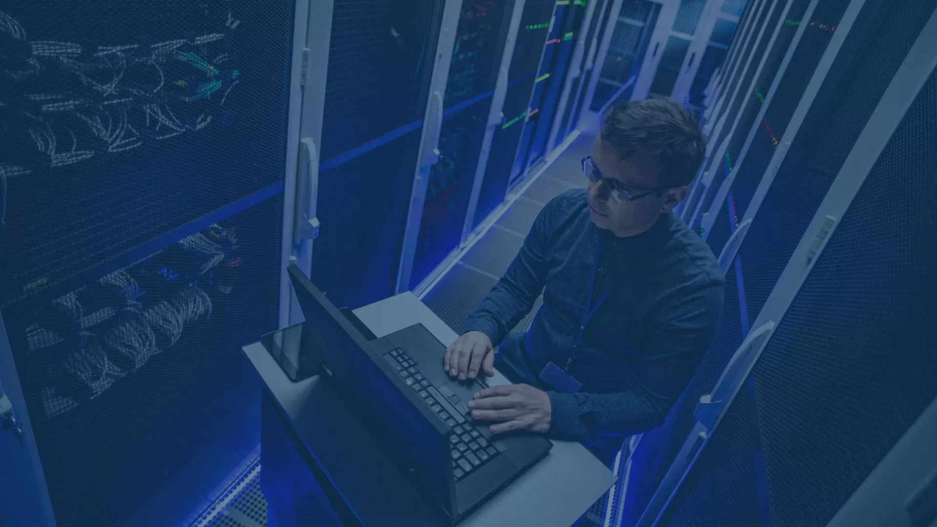 IT-Spezialist arbeitet im Rechenzentrum an einem Laptop, umgeben von Serverracks und blauer Beleuchtung.
