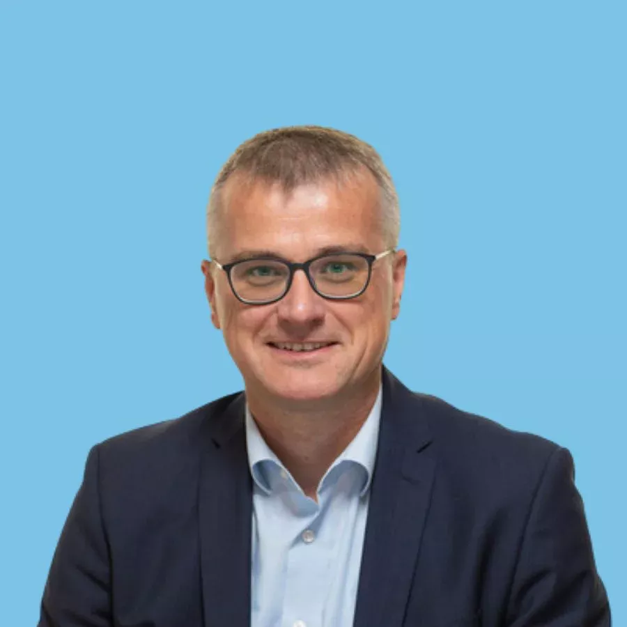 Porträtfoto eines lächelnden Mannes (Jürgen Hernadi) im Business-Outfit vor blauem Hintergrund.